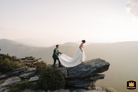 The groom carries the train of the bride’s dress as she steps across the rocky summit of Hawksbill Mountain during their sunset adventure session. 